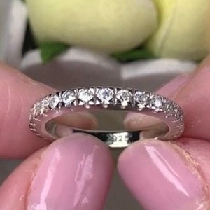 May include: A silver ring with a band of small, clear gemstones. The ring is held between two fingers with a soft pink nail polish. The background features yellow flowers.