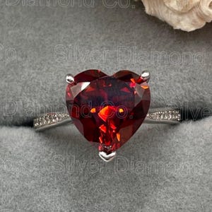 May include: A silver ring with a large heart-shaped red gemstone and smaller clear gemstones on the band.