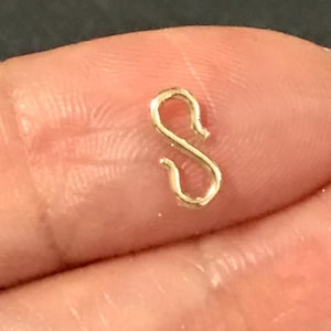 May include: A small, gold-colored S-shaped clasp. The clasp is held between two fingers, with a dark background. The clasp is likely used for jewelry making or as a decorative element.