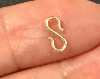 May include: A small, gold-colored S-shaped clasp. The clasp is held between two fingers, with a dark background. The clasp is likely used for jewelry making or as a decorative element.