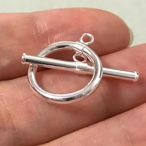 Könnte beinhalten: Ein glänzender silberner Knebelverschluss, bestehend aus einem kreisförmigen Ring und einem geraden Stab. Der Verschluss ist für die Schmuckherstellung konzipiert und wird vor einem neutralen Hintergrund gezeigt. Der Knebelverschluss ist ein gängiger Bestandteil von Halsketten und Armbändern.