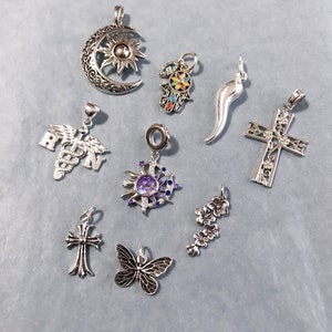 Puede incluir: Una colección de nueve colgantes de plata, que incluyen una luna creciente con un sol, una mano con un ojo, un cuerno, una cruz, un símbolo médico, un sol con una piedra morada, una mariposa, una flor y una cruz con un corazón.