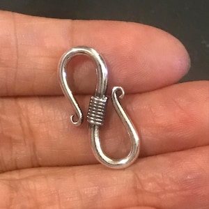 May include: A silver-colored hook clasp, featuring a curved design with a coiled center. The clasp has a smooth, polished finish and is likely used for jewelry or other small accessories. The clasp is held in a hand.