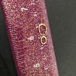 Puede incluir: Un par de pendientes de aro peque&ntilde;os, de color dorado. Los pendientes est&aacute;n colocados cerca de una regla rosa brillante, con medidas en cent&iacute;metros. Los pendientes son de dise&ntilde;o sencillo y parecen ser de metal.