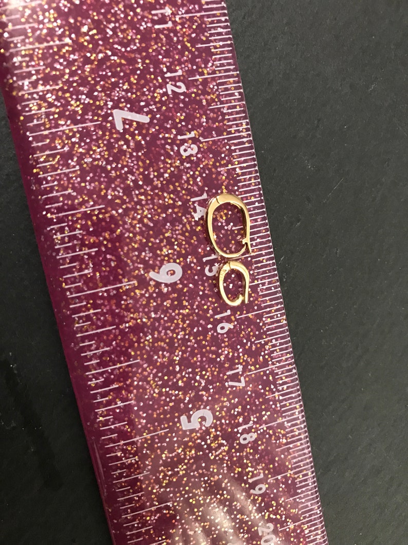 Puede incluir: Un par de pendientes de aro peque&ntilde;os, de color dorado. Los pendientes est&aacute;n colocados cerca de una regla rosa brillante, con medidas en cent&iacute;metros. Los pendientes son de dise&ntilde;o sencillo y parecen ser de metal.