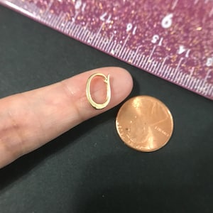 Puede incluir: Un peque&ntilde;o pendiente de color dorado en forma ovalada abierta. El pendiente se sostiene cerca de un centavo y una regla rosa con marcas negras. El pendiente es un dise&ntilde;o simple y elegante.