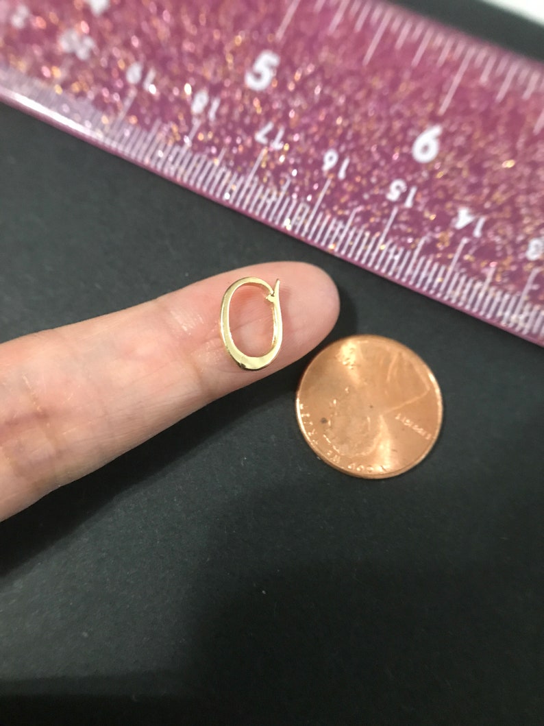 Puede incluir: Un peque&ntilde;o pendiente de color dorado en forma ovalada abierta. El pendiente se sostiene cerca de un centavo y una regla rosa con marcas negras. El pendiente es un dise&ntilde;o simple y elegante.