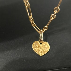 Puede incluir: Un collar dorado con un colgante en forma de coraz&oacute;n. El colgante presenta dise&ntilde;os grabados de s&iacute;mbolos y formas. La cadena tiene una mezcla de estilos de eslabones, con una combinaci&oacute;n de eslabones redondos y rectangulares peque&ntilde;os.