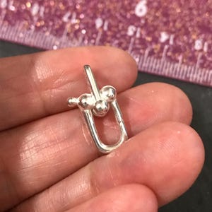 May include: A small, silver-colored jewelry clasp with a U-shaped base and a decorative top featuring three small spheres. The clasp is held in a person's hand, with a ruler in the background.