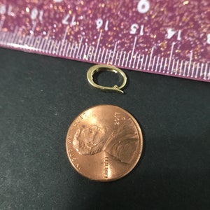 Puede incluir: Un peque&ntilde;o cierre de pendiente dorado junto a un centavo de cobre. El cierre del pendiente tiene forma ovalada con un cierre de bisagra. La moneda est&aacute; fechada en 2017. El fondo es una superficie oscura con una regla morada.