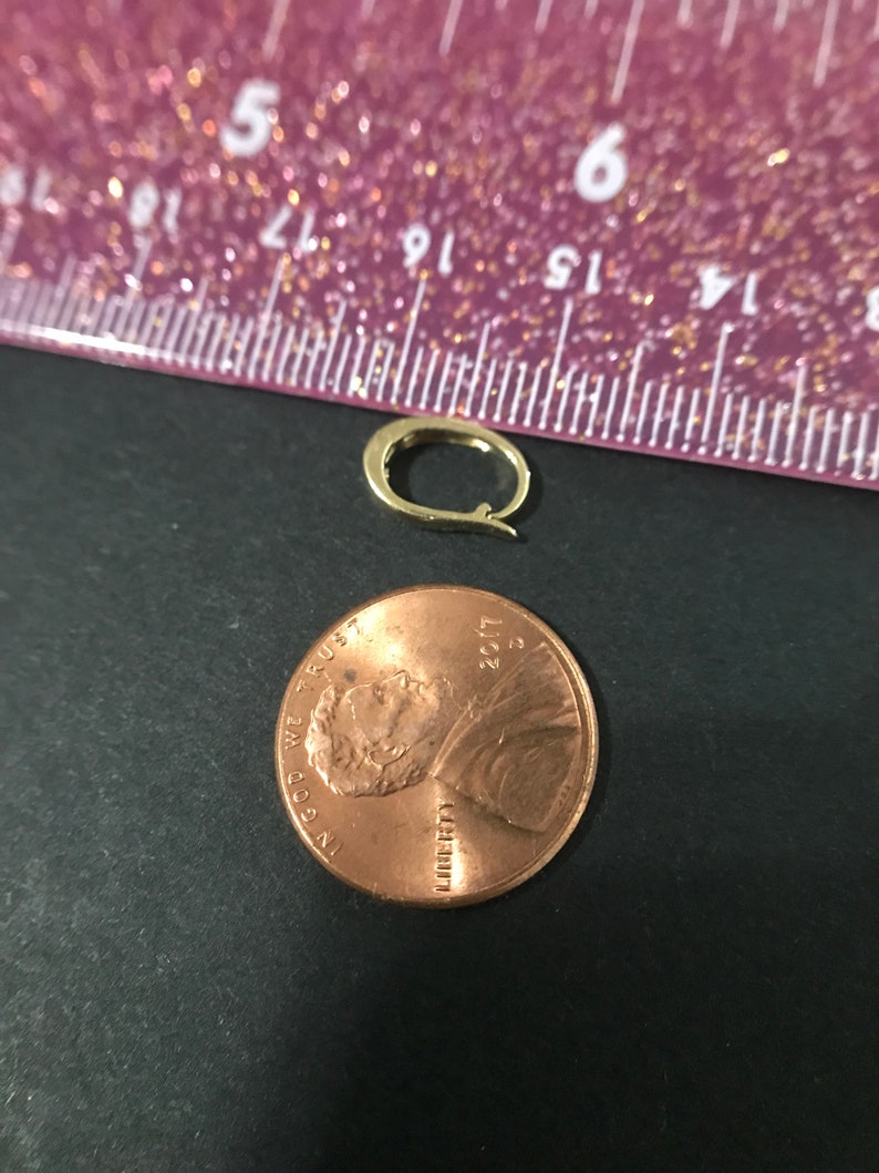 Puede incluir: Un peque&ntilde;o cierre de pendiente dorado junto a un centavo de cobre. El cierre del pendiente tiene forma ovalada con un cierre de bisagra. La moneda est&aacute; fechada en 2017. El fondo es una superficie oscura con una regla morada.