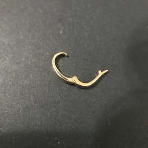 Puede incluir: Un solo pendiente de aro dorado, abierto y listo para ser abrochado. El pendiente es curvo y tiene un dise&ntilde;o simple y elegante. El fondo es de un color oscuro y s&oacute;lido.