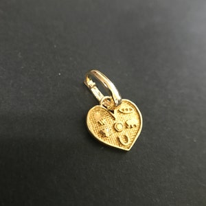 Puede incluir: Un colgante dorado en forma de coraz&oacute;n con un peque&ntilde;o aro. El colgante est&aacute; decorado con varios s&iacute;mbolos peque&ntilde;os, incluyendo un ojo, un elefante y una herradura. El colgante est&aacute; sobre un fondo oscuro.