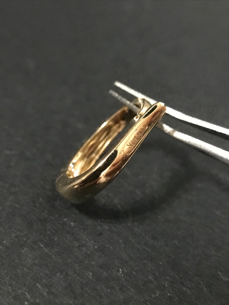 Puede incluir: Primer plano de un pendiente dorado de forma cuadrada. El pendiente est&aacute; sujeto con unas pinzas sobre un fondo oscuro. El texto "G14K" es visible en el pendiente.