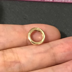 May include: A small, gold-colored circular clasp, likely for jewelry. The clasp is open, revealing a small hinge. The background is a hand holding the clasp, with a ruler in the background.