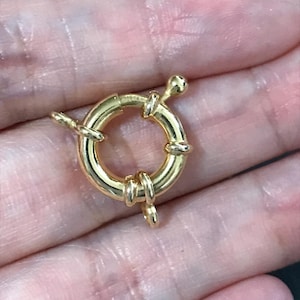 Può includere: Primo piano di una chiusura per gioielli rotonda color oro. La chiusura ha un design ad anello a molla con un piccolo pomello arrotondato su un'estremità e un anello sull'altra. La chiusura è progettata per fissare i gioielli.