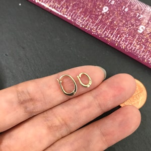 Puede incluir: Dos peque&ntilde;os pendientes de aro dorados. Uno es ligeramente m&aacute;s grande que el otro. Los pendientes est&aacute;n en la mano de una persona, junto a un centavo. Una regla rosa con purpurina dorada est&aacute; en el fondo.