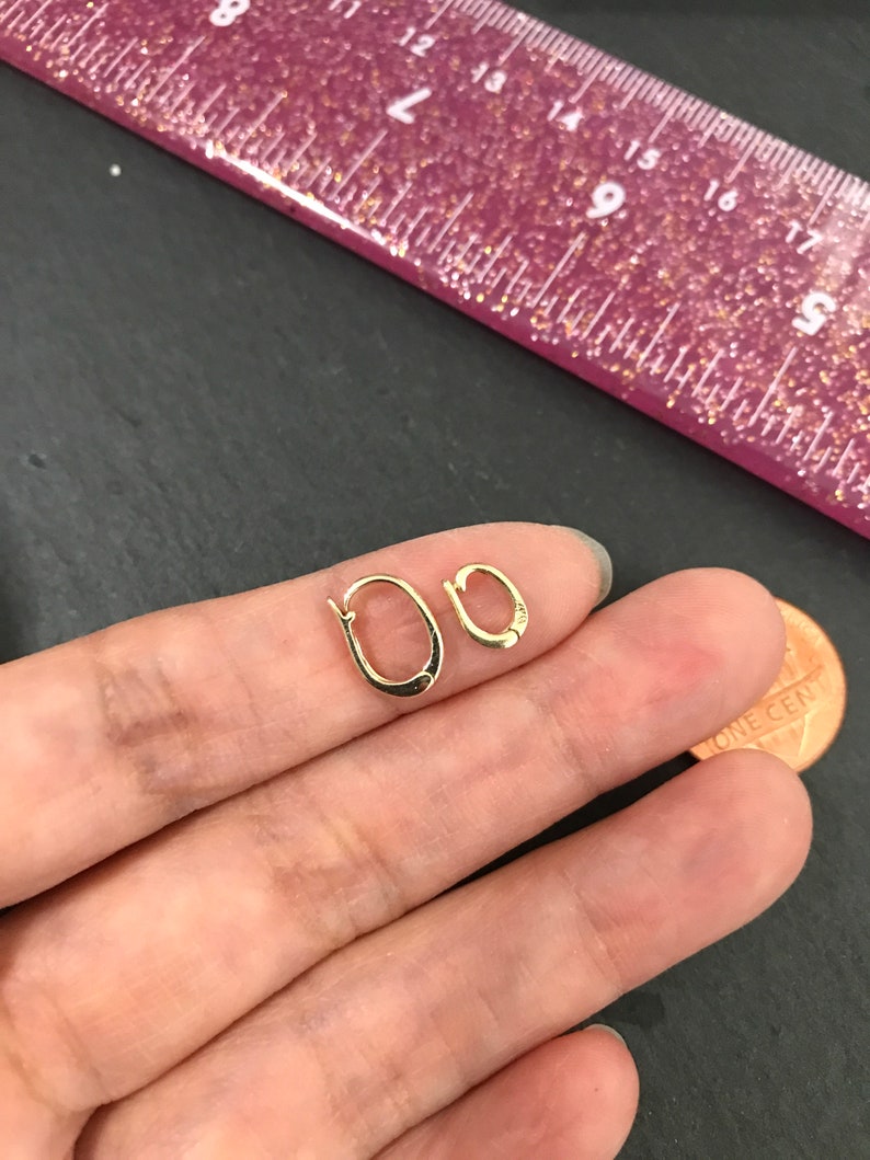 Puede incluir: Dos peque&ntilde;os pendientes de aro dorados. Uno es ligeramente m&aacute;s grande que el otro. Los pendientes est&aacute;n en la mano de una persona, junto a un centavo. Una regla rosa con purpurina dorada est&aacute; en el fondo.