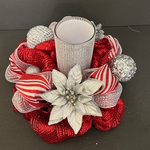May include: A festive centerpiece featuring a clear glass candle holder surrounded by red and white striped ribbon, silver ornaments, and a silver poinsettia. The candle holder is embellished with silver rhinestones, creating a sparkling holiday decoration.