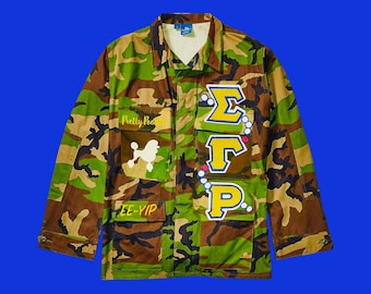 Pode incluir: Uma jaqueta de camuflagem com uma variedade de patches, incluindo um poodle branco, um patch amarelo "Pretty Poodles" e um patch azul e amarelo com as letras "ΣΦΡ".