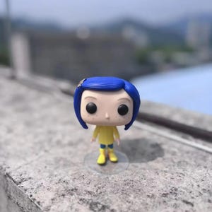 May include: A vinyl figure of a character with blue hair, wearing a yellow outfit and blue boots. The figure has large black eyes and is standing on a clear plastic base. A small decorative hair clip is visible.