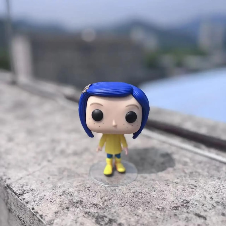 May include: A vinyl figure of a character with blue hair, wearing a yellow outfit and blue boots. The figure has large black eyes and is standing on a clear plastic base. A small decorative hair clip is visible.
