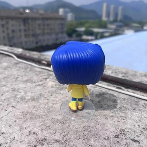 May include: A vinyl figure of a character with blue hair, wearing a yellow jacket, blue pants, and yellow boots. The figure is standing on a clear plastic stand. The background shows a cityscape and mountains.