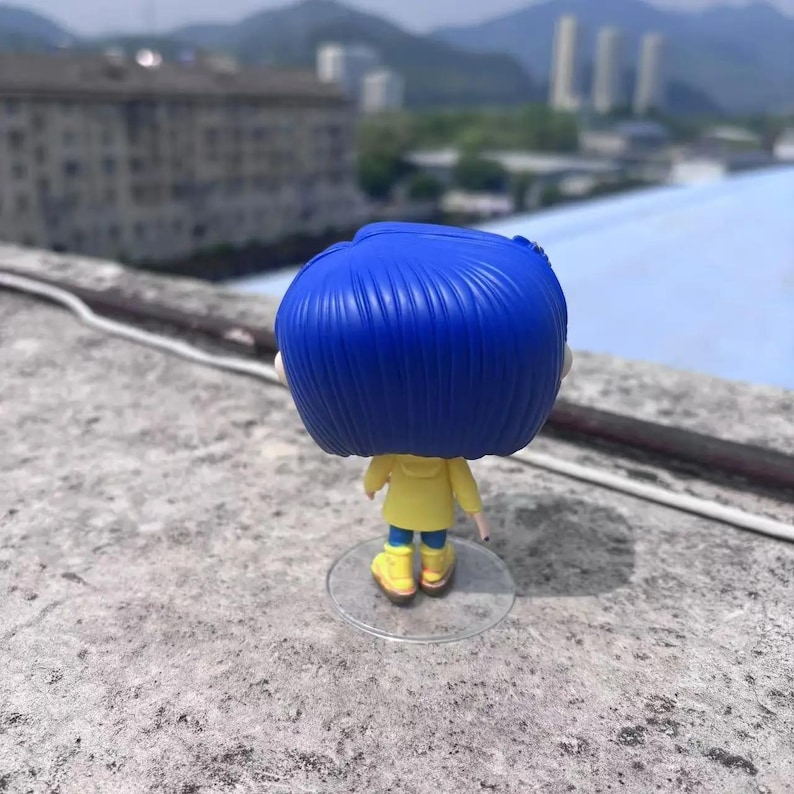 May include: A vinyl figure of a character with blue hair, wearing a yellow jacket, blue pants, and yellow boots. The figure is standing on a clear plastic stand. The background shows a cityscape and mountains.
