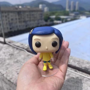 May include: A vinyl figure of a character with blue hair, black eyes, and freckles, wearing a yellow jacket and boots. The figure is holding a small object and is on a clear stand.