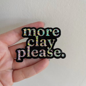 May include: A black sticker with iridescent glitter lettering that reads "more clay please."