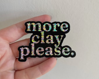 Könnte beinhalten: Ein schwarzer Aufkleber mit schimmerndem Glitzer-Schriftzug, der "more clay please." lautet.