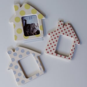 May include: Three house-shaped picture frames with polka dot patterns. One frame is white with yellow dots and contains a photograph of a cat. Another frame is white with red dots, and the third frame is white with light blue dots.