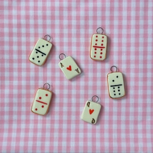 May include: A collection of small, rectangular charms featuring domino and playing card designs. The charms are cream-colored with black and red markings, and gold-colored edges. Each charm has a small metal loop at the top. The background is a pink and white checkered pattern.