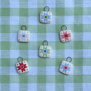 May include: Six square ceramic pendants with black metal loops. Each pendant features a colorful starburst design with a heart in the center. The color schemes include red, blue, green, and purple on a cream background. The pendants are arranged on a green and white checkered fabric.