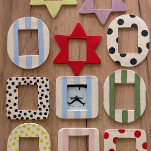 May include: A collection of eleven ceramic picture frames in various shapes including stars, circles, and squares. The frames feature diverse patterns such as blue stripes, black polka dots, green stripes, and red dots, with one frame containing a photo.