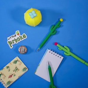 May include: A flat lay of a blue background with a yellow cactus-shaped stress ball, a notepad with a spiral binding, two green cactus-shaped pens, and a sticker that says "Little Pricks".