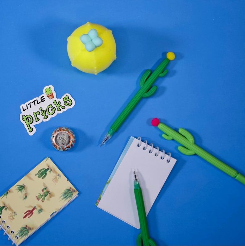 May include: A flat lay of a blue background with a yellow cactus-shaped stress ball, a notepad with a spiral binding, two green cactus-shaped pens, and a sticker that says "Little Pricks".