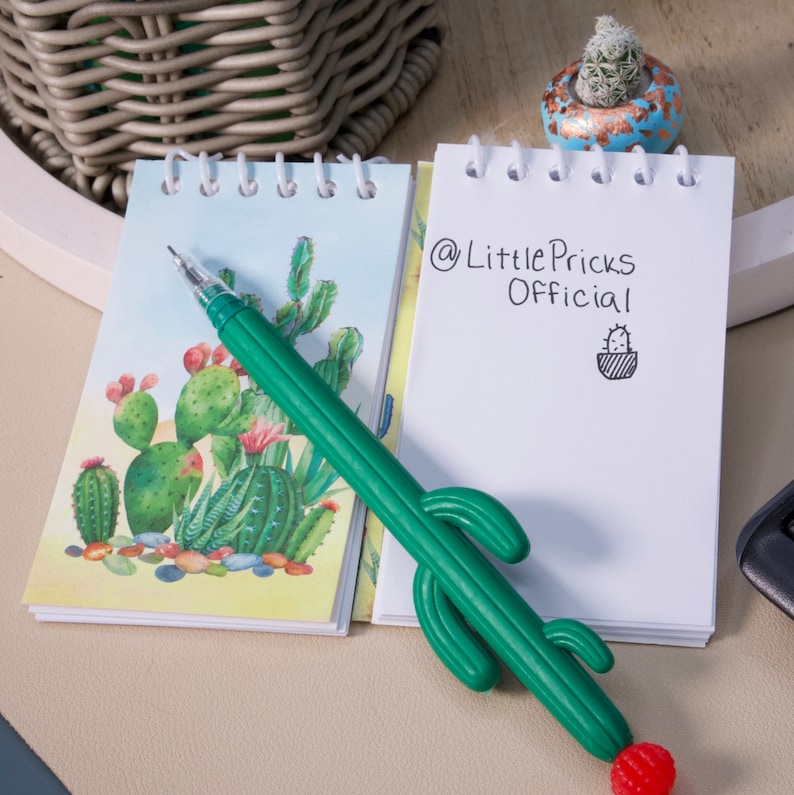 May include: Two small spiral bound notebooks with cactus illustrations. One notebook has a green cactus pen with a red flower on the end. The other notebook has the text "@LittlePricks Official" written on it.
