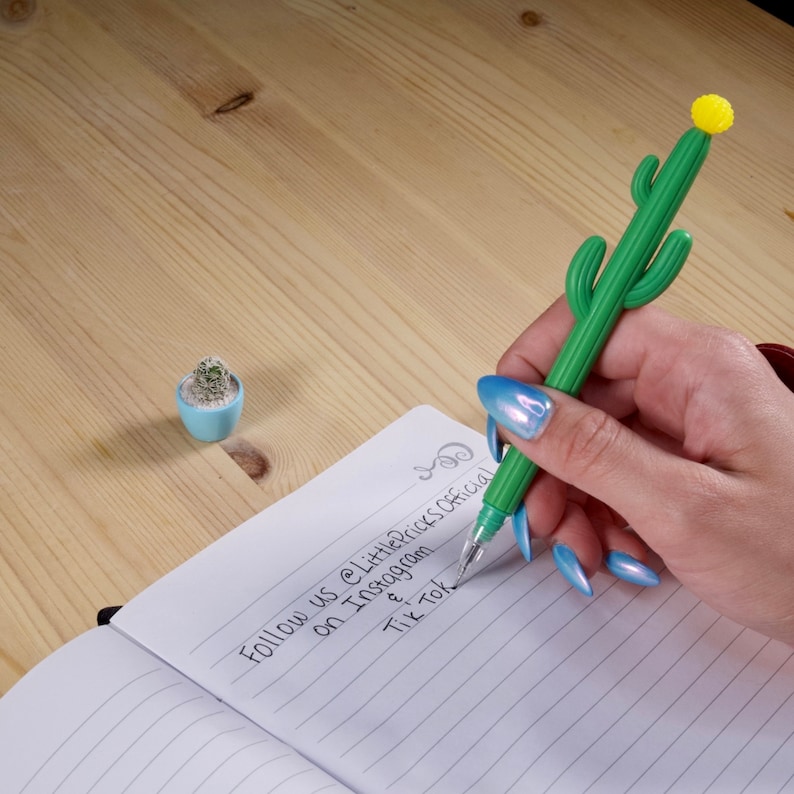 May include: A hand holding a green cactus-shaped pen with a yellow flower on top. The pen is writing in a notebook with the text "Follow us @LittlePriceOfficial on Instagram & TikTok".