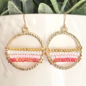 May include: Gold hoop earrings with a multi-coloured beaded design. The beads are pink, white, and gold, and they are arranged in a horizontal stripe pattern. The earrings are a simple and elegant design that would be perfect for any occasion.