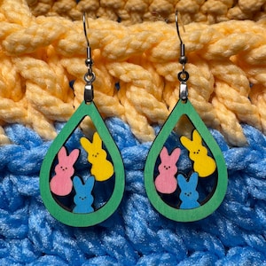 May include: A pair of green teardrop-shaped earrings with three colorful bunny-shaped cutouts. The bunnies are pink, yellow, and blue.
