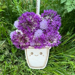 Può includere: Un vaso di legno bianco con un viso sorridente dipinto, pieno di un mazzo di fiori viola e bianchi.