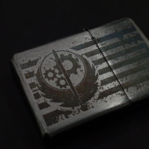 May include: A metallic grey lighter with a distressed finish. The lighter features a design with a circular emblem, gears, a sword, and striped pattern. The design is etched into the metal, creating a vintage look.