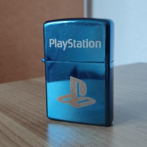 Puede incluir: Un encendedor de metal azul con la palabra "PlayStation" y el logotipo de PlayStation grabado en la parte delantera.