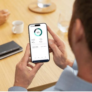 以下が含まれることがあります： 金融ダッシュボードを表示するスマートフォンを手に持っている人。円形の進捗インジケーターは100％です。画面にはデータと「Efficiency Pulse」のテキストが表示されます。スマートフォンは木製のテーブルの上に置かれています。