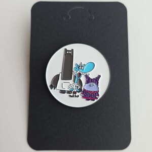 May include: A round enamel pin featuring a white background with three cartoon characters: a tall dark gray figure, a light blue creature with a horn, and a small purple character with dark hair, all mounted on a rectangular black display card.