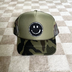 May include: A green and black camouflage trucker hat with a black smiley face embroidered on the front.