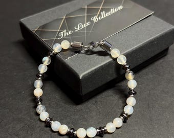 Puede incluir: Un brazalete de cuentas blancas y negras con un cierre plateado. El brazalete está en una caja negra con el texto "The Lux Collection" en la tapa.