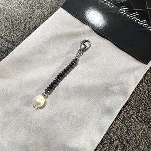 Puede incluir: Una cadena de cuentas negras con un dije de perla blanca y un cierre plateado. La cadena está sobre un fondo de tela blanca con el texto "The Lux Collection" visible en el fondo.