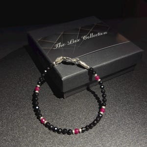 May include: A black and red beaded bracelet with a silver clasp. The bracelet is on a black surface with a box that says "The Lux Collection".